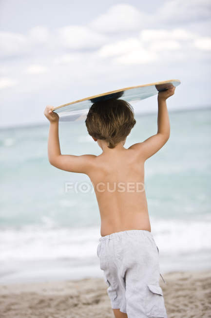 Rückansicht eines kleinen Jungen, der am Strand ein Brett über dem Kopf hält — Stockfoto