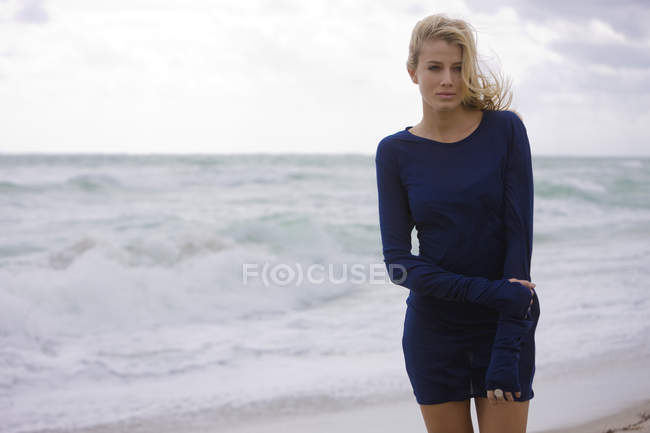 Задумчивая блондинка в платье, стоящая на ветреном пляже — стоковое фото
