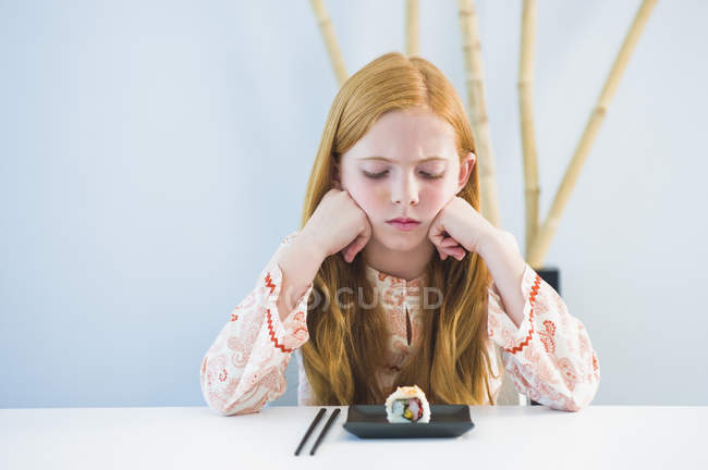 Грустная рыжая девушка сидит за обеденным столом и смотрит на суши — стоковое фото