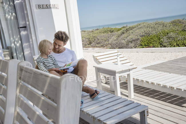 Счастливые отец и сын читают книгу на крыльце прибрежного дома — стоковое фото