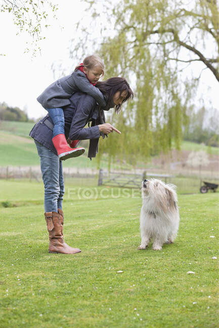 Жінка, що носить доньку на спині і лоскотає свого собаку — стокове фото