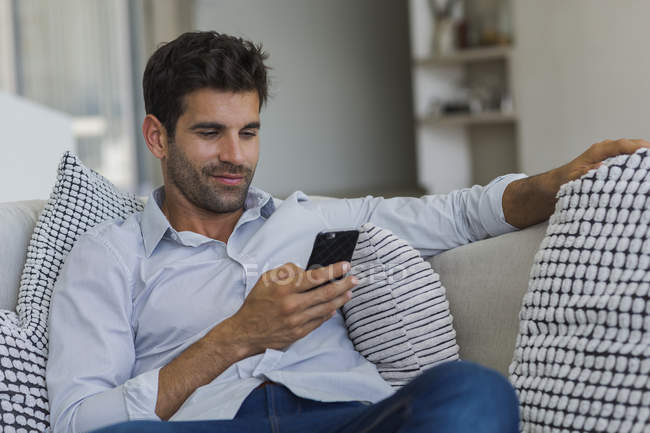 Uomo sorridente che usa il cellulare sul divano di casa — Foto stock