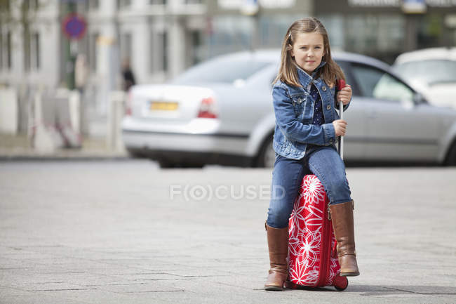 Portrait of little girl in denim clothes sitting on luggage on street — Stock Photo