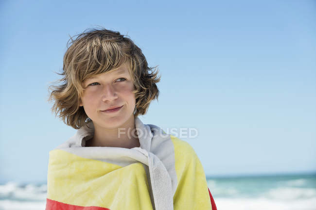 Thoughtful teenage boy wrapped in towel standing on beach — Stock Photo