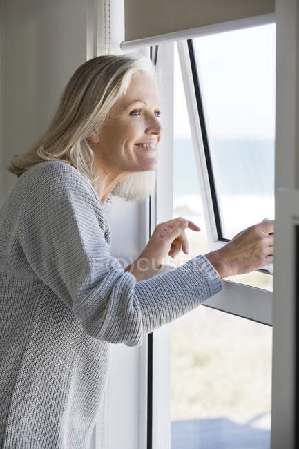 Sorrindo mulher sênior olhando através da janela na casa — Fotografia de Stock