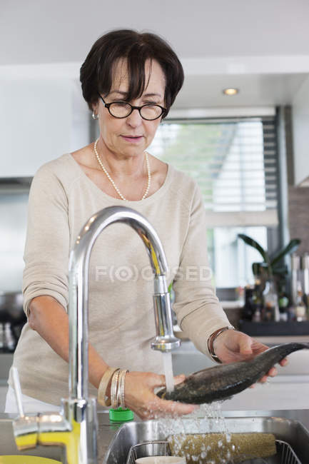 Mujer mayor lavando pescado en fregadero de cocina - foto de stock