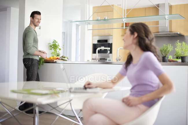 Woman using laptop with husband preparing food on background — Stock Photo