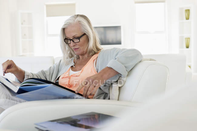 Mulher sênior sentado no sofá e livro de leitura em casa — Fotografia de Stock