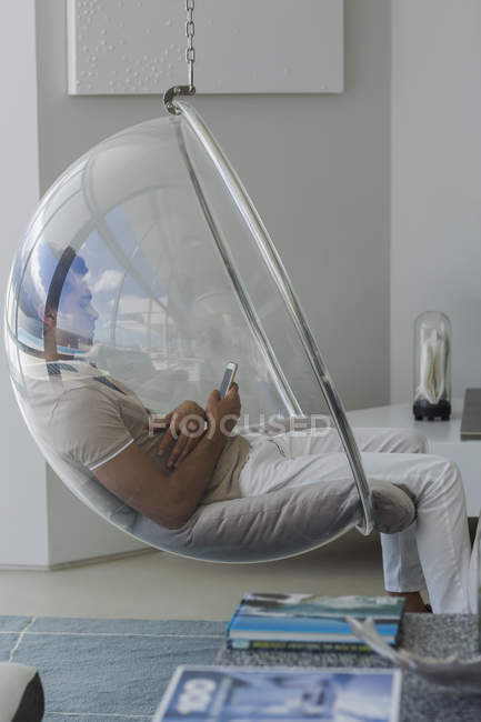 Молодой человек сидит в стеклянном кресле и пользуется мобильным телефоном дома — стоковое фото