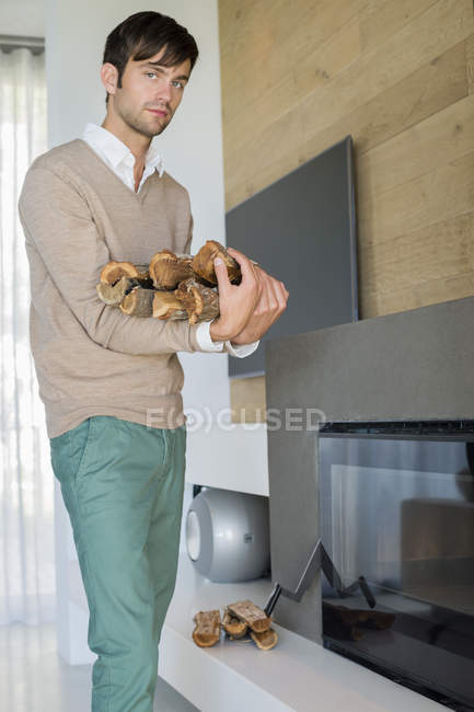 Retrato de un joven llevando leña cerca de la chimenea en la sala de estar - foto de stock
