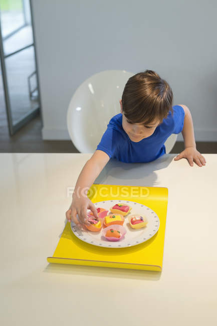 Niño recogiendo un cupcake del plato en la mesa de la cocina - foto de stock