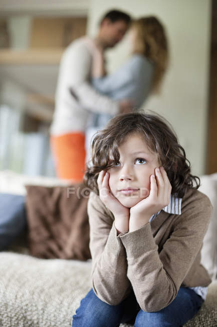 Petit garçon réfléchi assis sur le canapé avec les parents romancer sur le fond — Photo de stock