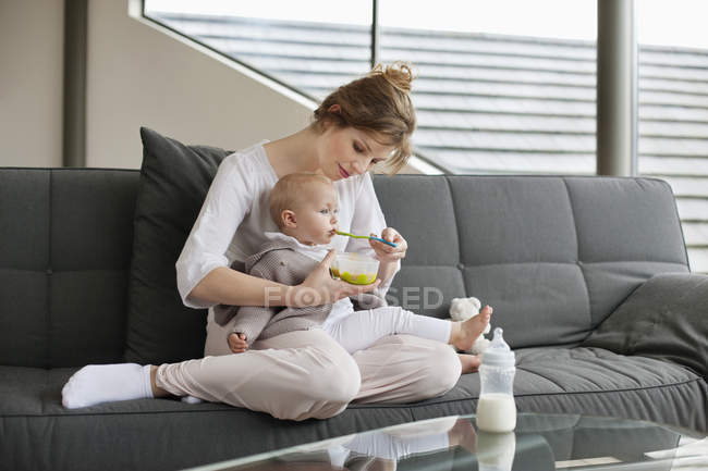 Mulher alimentando bebê filha no sofá em casa — Fotografia de Stock