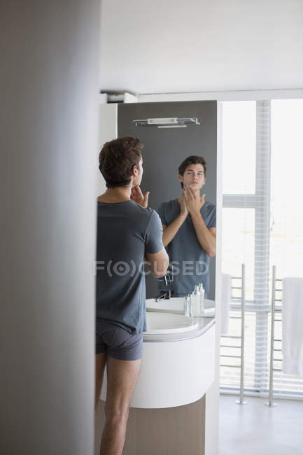 Молодой человек надевает лосьон после бритья на лицо перед зеркалом в ванной — стоковое фото