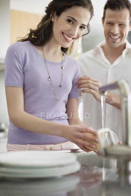Woman washing utensils in kitchen with husband on background — Stock Photo
