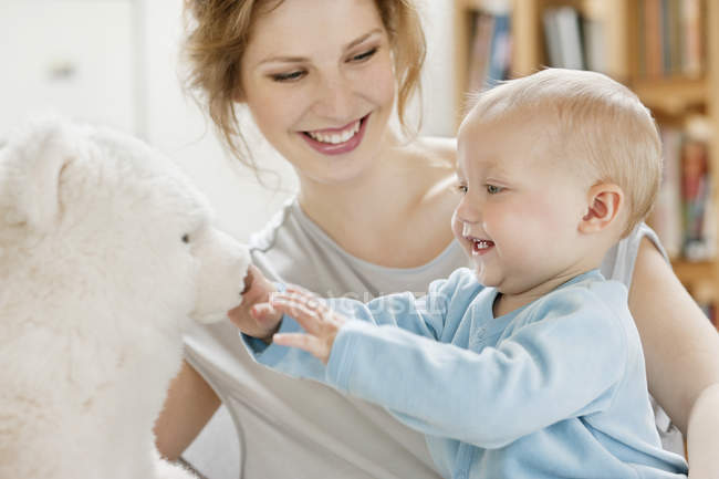 Baby girl playing with teddy bear and laughing with mother on background — Stock Photo