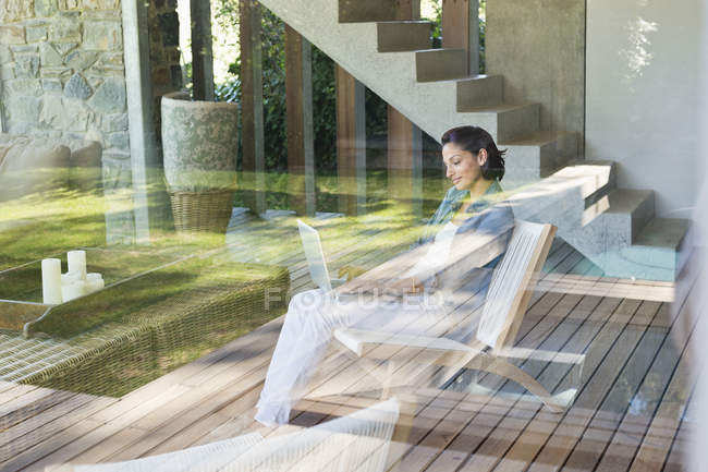 Mulher usando laptop na poltrona em casa visto através do painel da janela — Fotografia de Stock