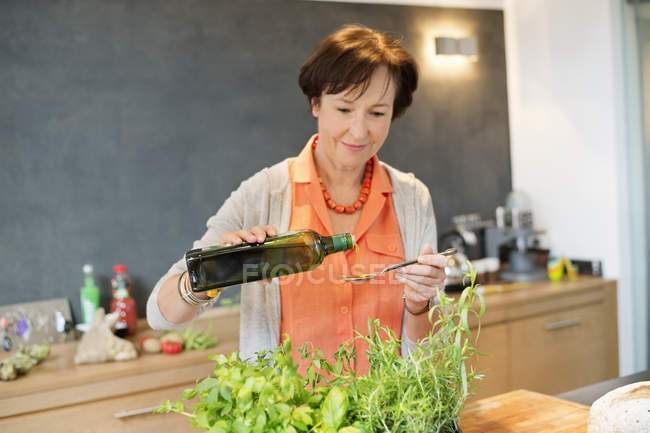 Senior woman pouring olive oil into spoon in kitchen — Stock Photo