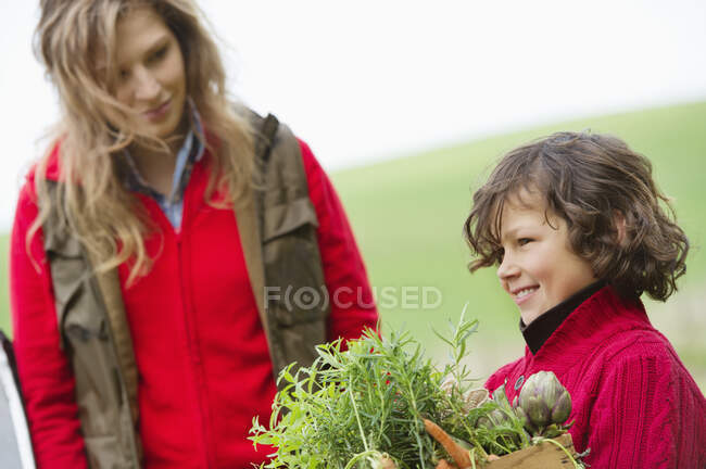 Garçon tenant une caisse de légumes assortis avec sa mère dans une ferme — Photo de stock