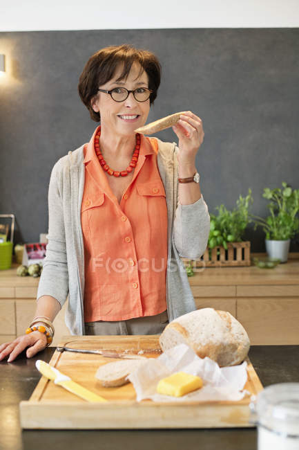 Портрет усміхненої старшої жінки, що їсть хліб на кухні — стокове фото