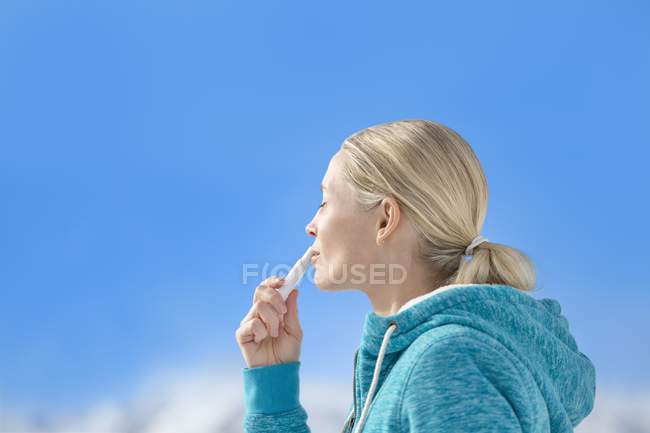 Mujer rubia aplicando bálsamo labial en los labios contra el cielo azul - foto de stock