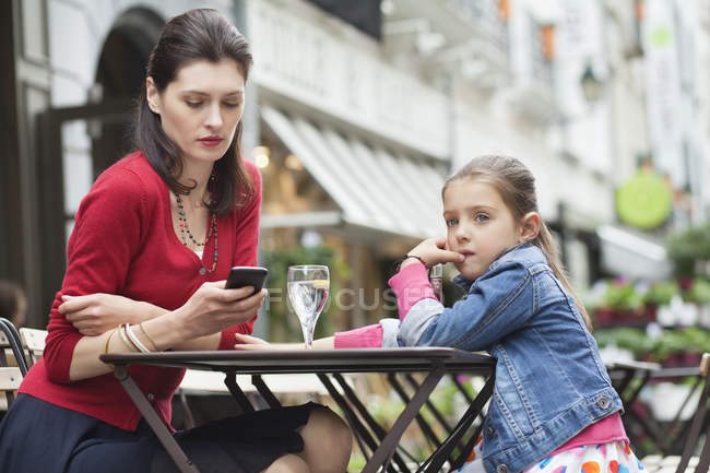 Mujer con hija pequeña sentada en un café y usando teléfono móvil - foto de stock