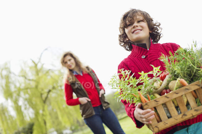 Garçon tenant une caisse de légumes assortis avec sa mère dans une ferme — Photo de stock