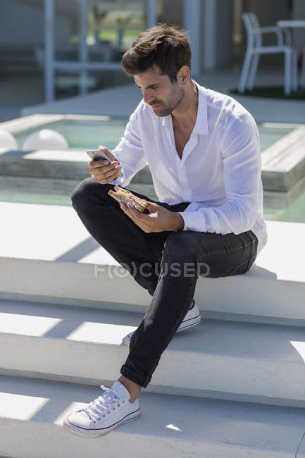 Confident man using smartphone and eating sandwich on stairs outdoors — Stock Photo