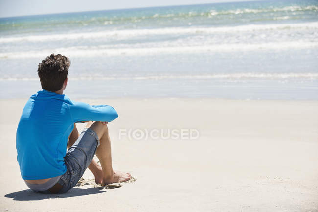 Hombre de ensueño sentado en la playa de arena y mirando a la vista - foto de stock