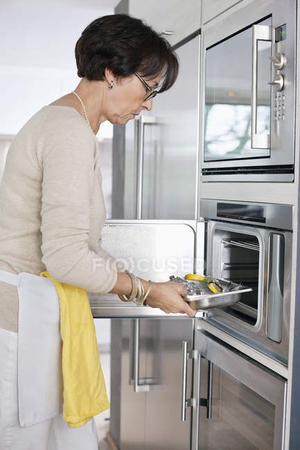 Mulher sênior colocando bandeja de frutos do mar no forno na cozinha — Fotografia de Stock