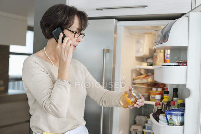 Senior woman looking at a refrigerator and talking on mobile phone in kitchen — Stock Photo