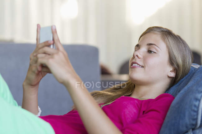 Улыбающаяся девочка-подросток лежит на бобовом мешке и пользуется мобильным телефоном — стоковое фото