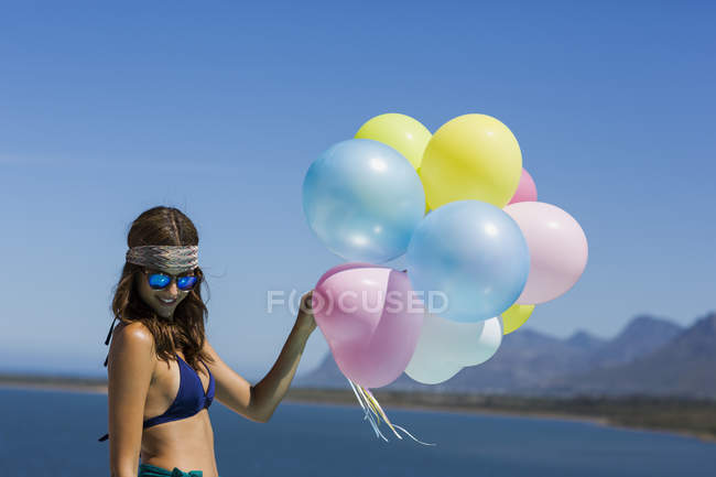 Elegante jovem segurando balões coloridos na margem do lago contra o céu azul — Fotografia de Stock
