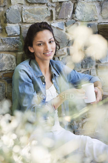 Portrait of smiling woman leaning against brick wall with cup of coffee — Stock Photo