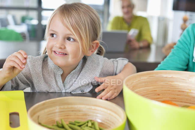Nahaufnahme eines lächelnden süßen kleinen Mädchens, das mit Schalen voller Gemüse am Tisch sitzt — Stockfoto