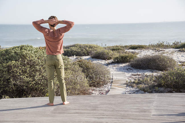 Junger Mann steht auf Holzterrasse und blickt aufs Meer — Stockfoto