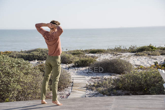 Молодой человек стоит на деревянной террасе и смотрит на море — стоковое фото