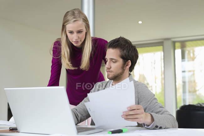 Mulher olhando para o homem trabalhando no laptop e segurando documentos — Fotografia de Stock