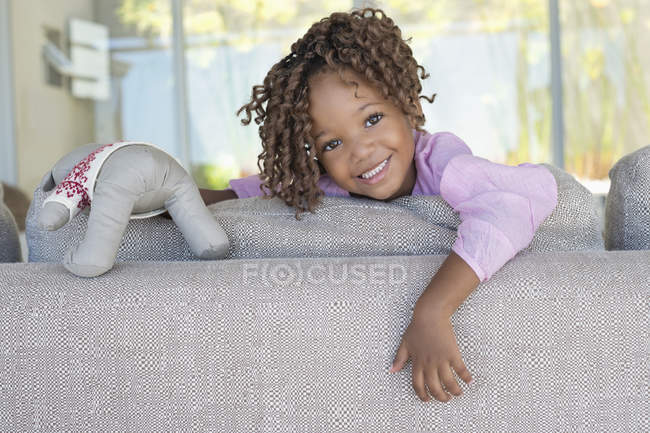 Portrait of smiling little girl holding teddy bear on sofa in room — Stock Photo