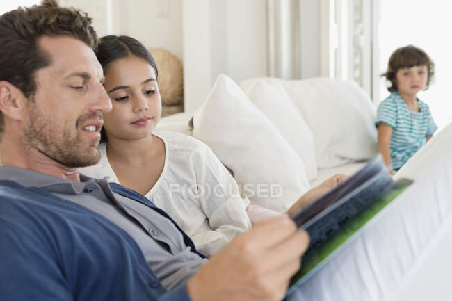 Hombre leyendo una revista con su hija y su hijo en el fondo - foto de stock