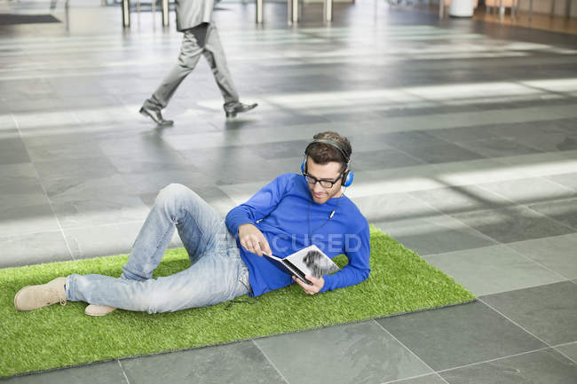 Empresário relaxante na grama falsa, ouvindo música e lendo livro no lobby do escritório — Fotografia de Stock
