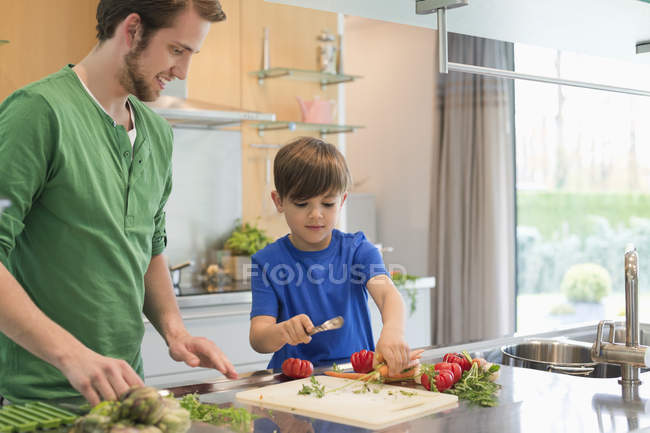 Чоловік дивиться на сина, який ріже овочі на кухні — стокове фото
