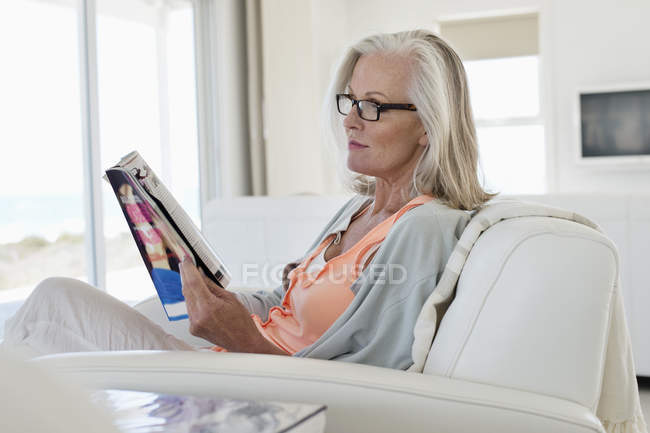 Mulher sentada no sofá e revista de leitura em casa — Fotografia de Stock
