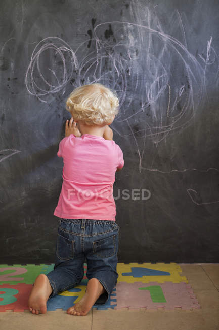 Rear view of cute blonde little girl writing on a blackboard — Stock Photo