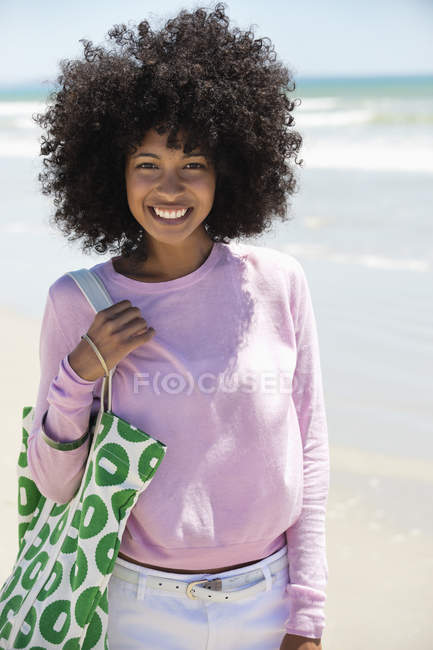 Портрет улыбающейся молодой женщины с узорчатой сумкой, стоящей на пляже — стоковое фото