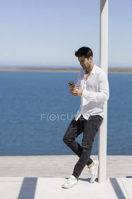Уверенный в себе человек, опираясь на полюс на берегу озера и используя смартфон — стоковое фото