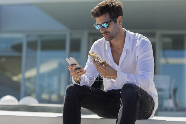 Selbstbewusster Mann nutzt Smartphone und isst Sandwich im Freien — Stockfoto