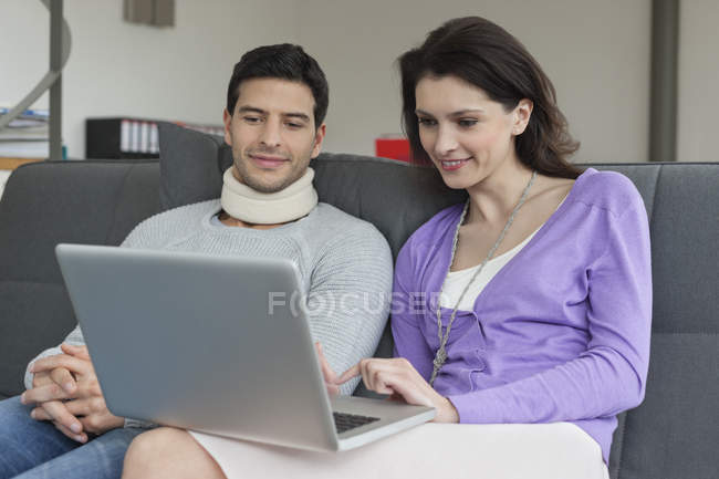 Женщина с ноутбуком и мужем с болями в шее сидит на диване — стоковое фото