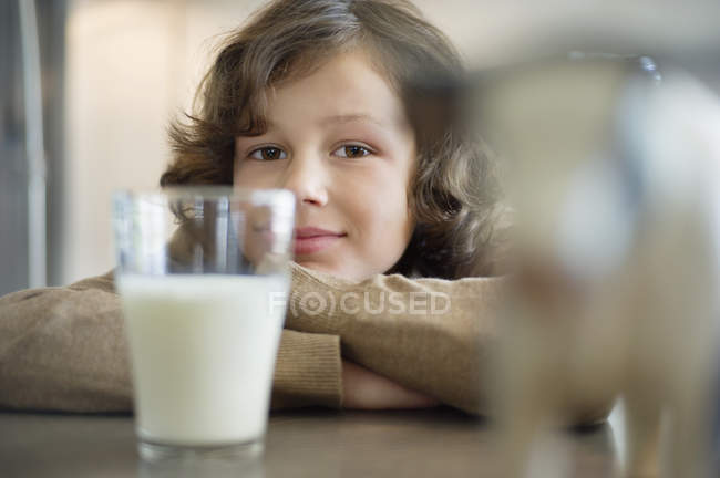 Retrato de menino sorridente inclinado na mesa com copo de leite — Fotografia de Stock