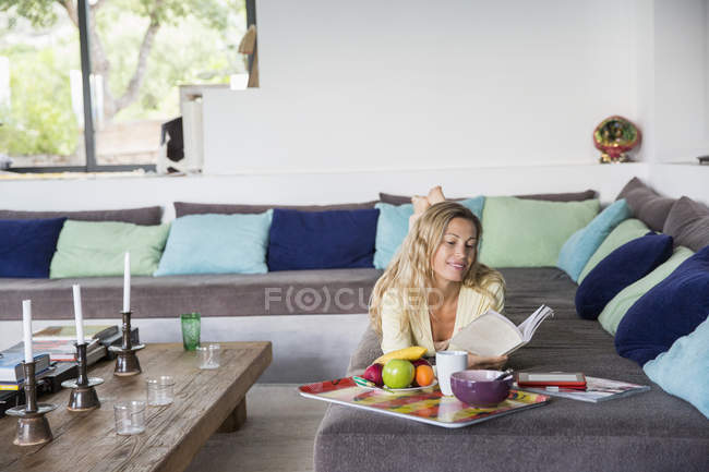 Attractive woman reading book while lying on sofa with tray of food in hotel — Stock Photo